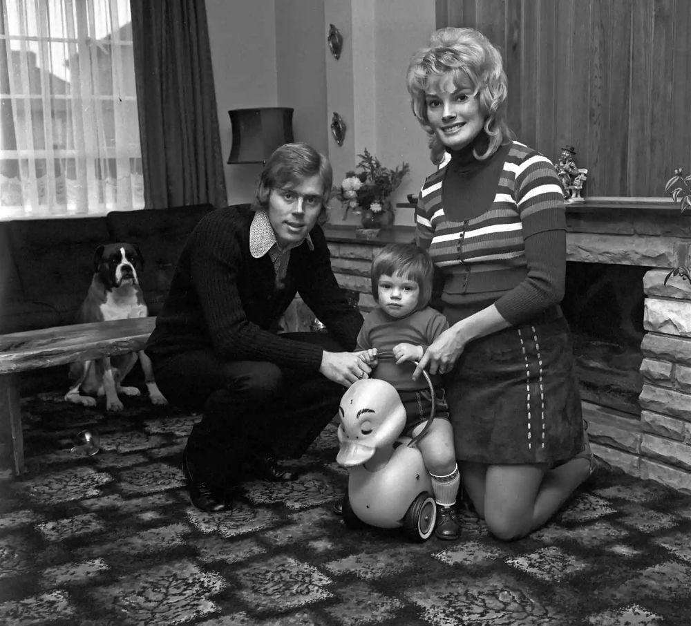 A man and woman pose with a young child sitting on a duck-shaped toy in a living room. A dog sits on a bench in the background. The room has patterned carpet, a stone fireplace, and curtains letting in natural light.