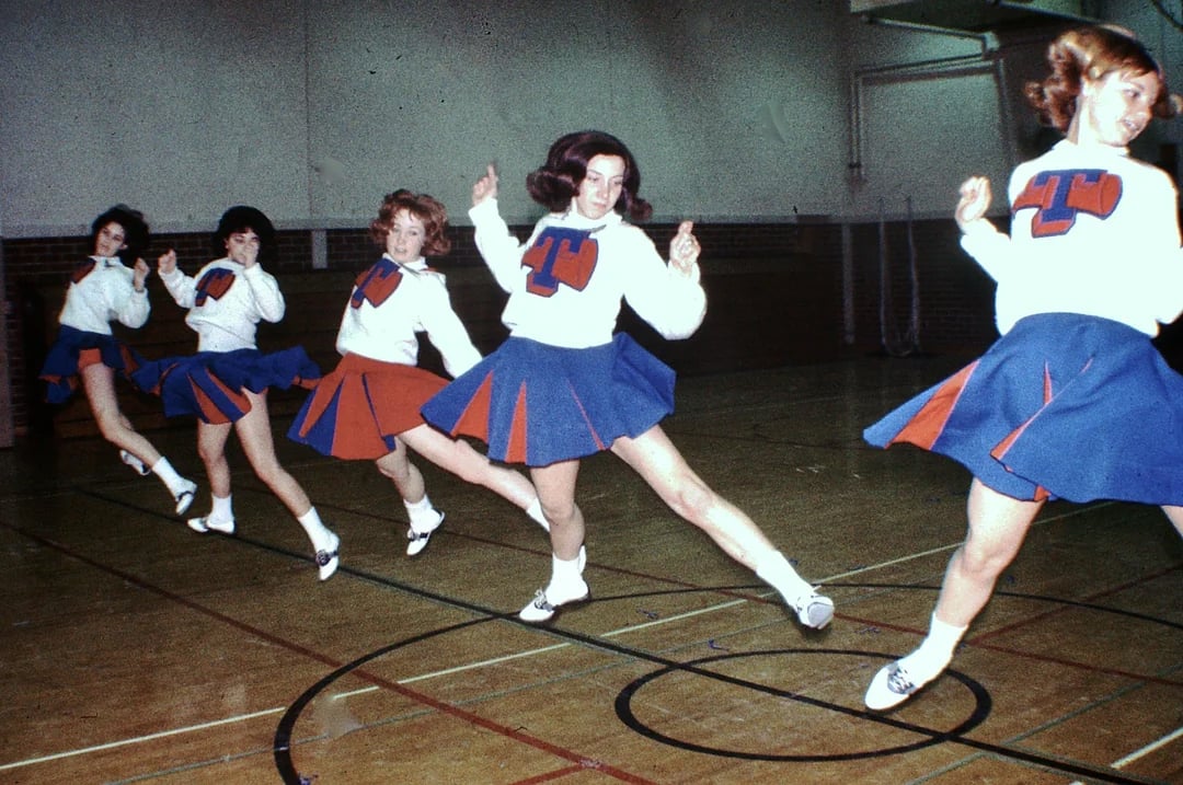 Five cheerleaders in matching blue and orange uniforms perform a synchronized high kick routine inside a gymnasium with wooden floors and brick walls.