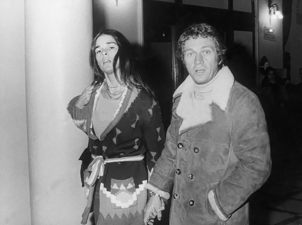 A man and woman walk hand in hand at night, dressed in 1970s-style clothing. The man wears a shearling coat and turtleneck, while the woman wears a patterned dress with a belt. They appear to be entering a building.