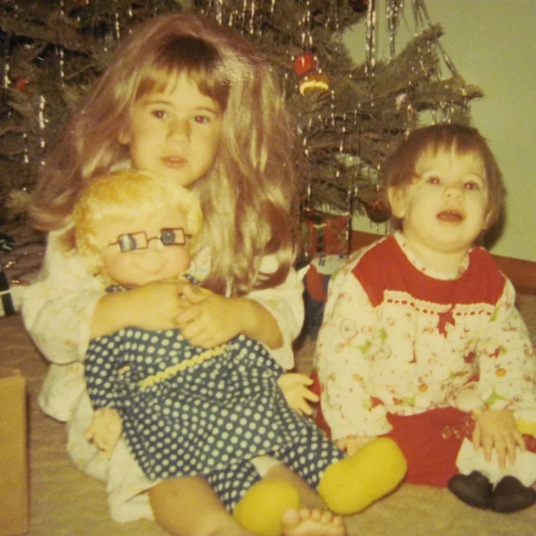 Two young children sit in front of a decorated Christmas tree. The older child holds a large doll with glasses and blonde hair, while the younger child, wearing a white and red outfit, sits beside them and smiles.