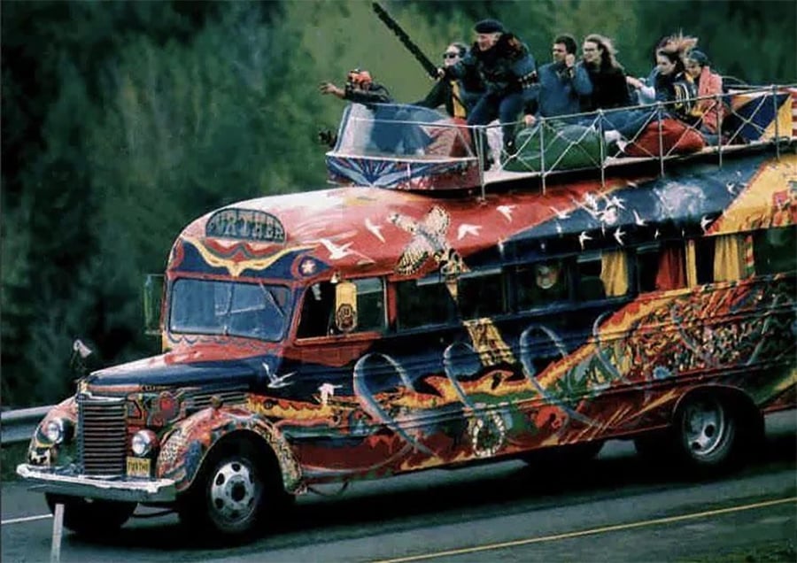 A colorfully painted, psychedelic bus drives down a road with several people riding on the roof, some sitting and others standing, surrounded by trees in the background.