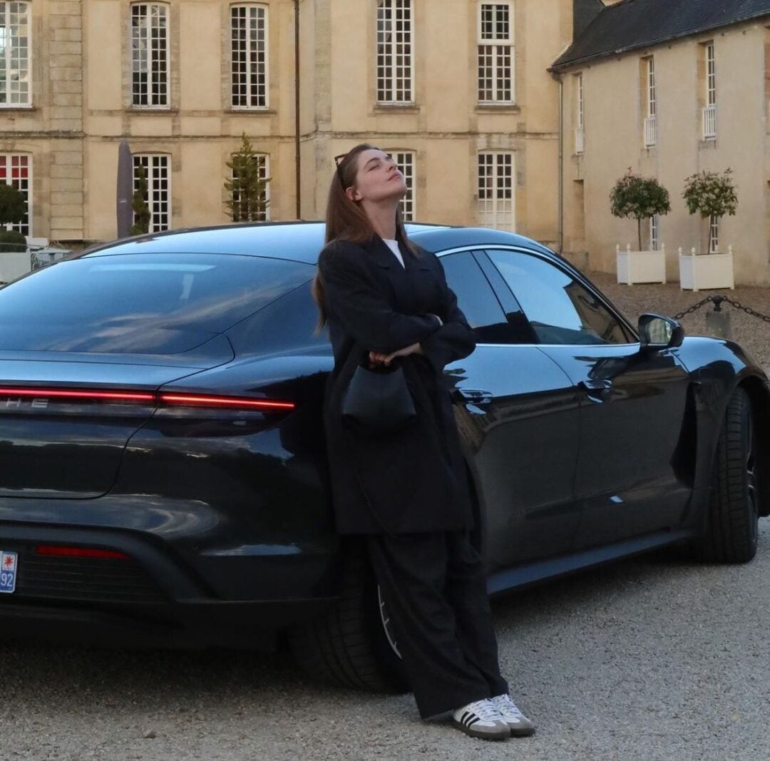 A woman in a dark coat and sneakers leans against the back of a sleek black car, arms crossed and head tilted up, in front of a building with tall windows and cream-colored walls.