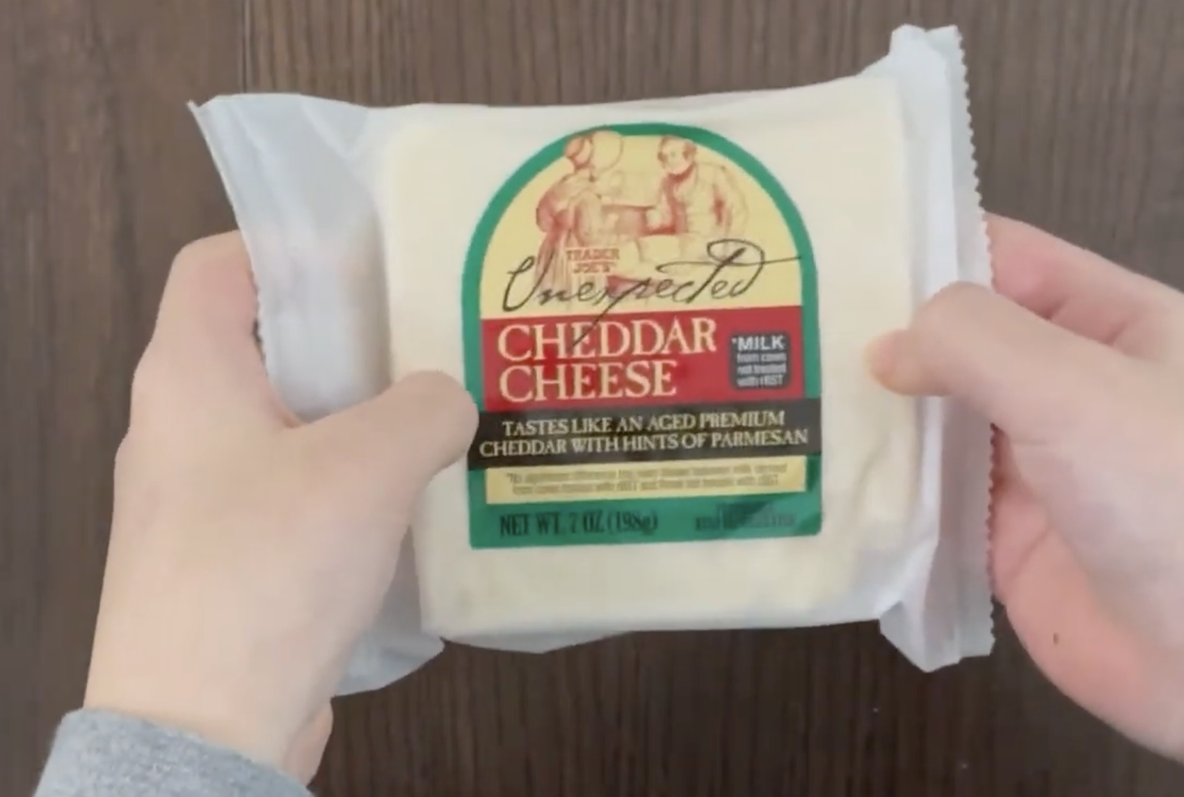 Two hands holding a block of Trader Joe’s Unexpected Cheddar Cheese in its plastic packaging.