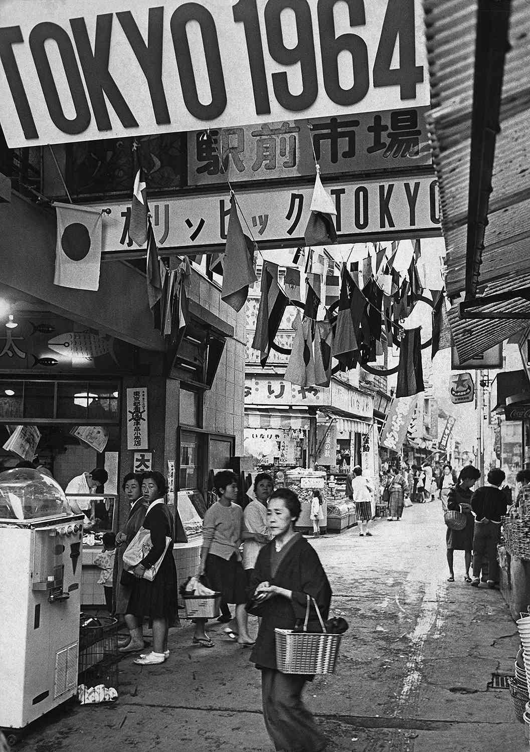 A busy street scene in Japan during the 1964 Tokyo Olympics, with crowds of people, hanging flags, market shops, and large banners displaying “TOKYO 1964” overhead.