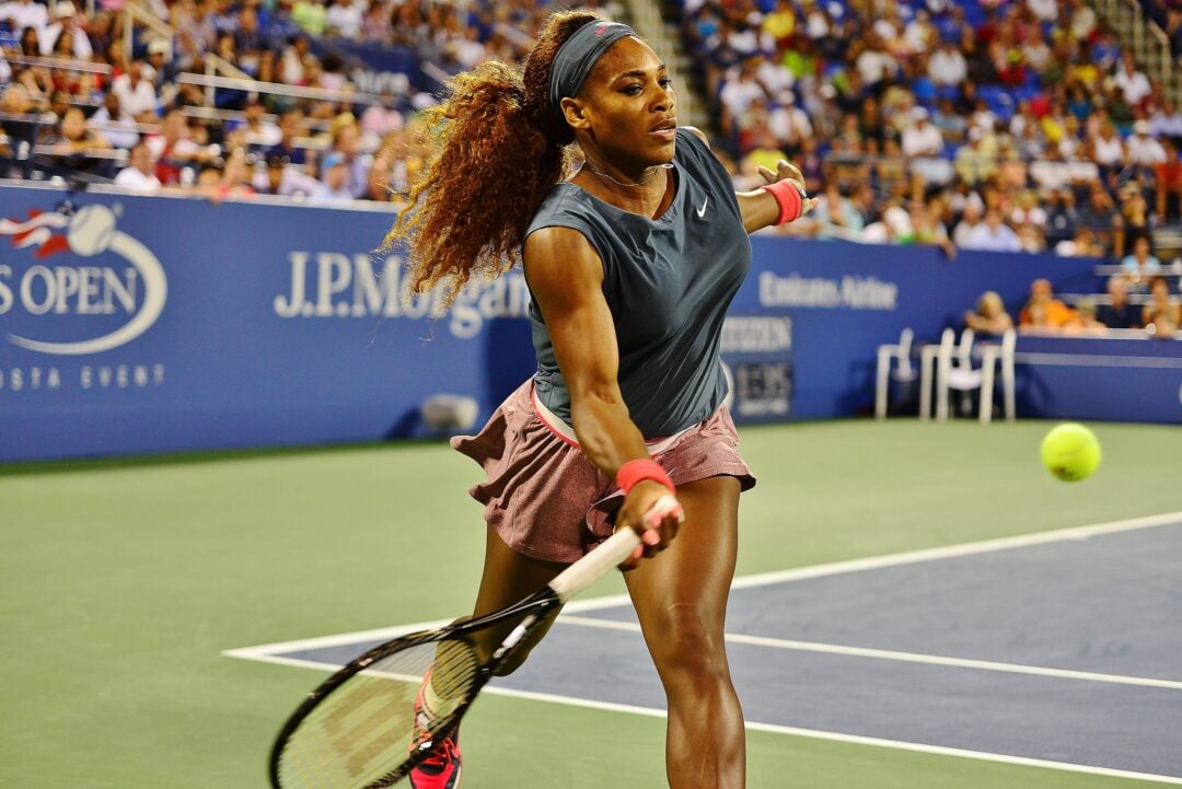 A female tennis player in athletic wear prepares to hit a tennis ball with a forehand swing on a hard court during a professional match, with spectators watching in the background.