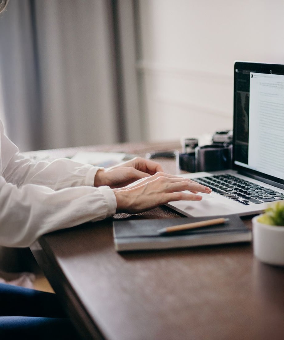 A person in a white shirt types on a laptop at a wooden desk, with notebooks, a pencil, camera lenses, and a small potted plant nearby.