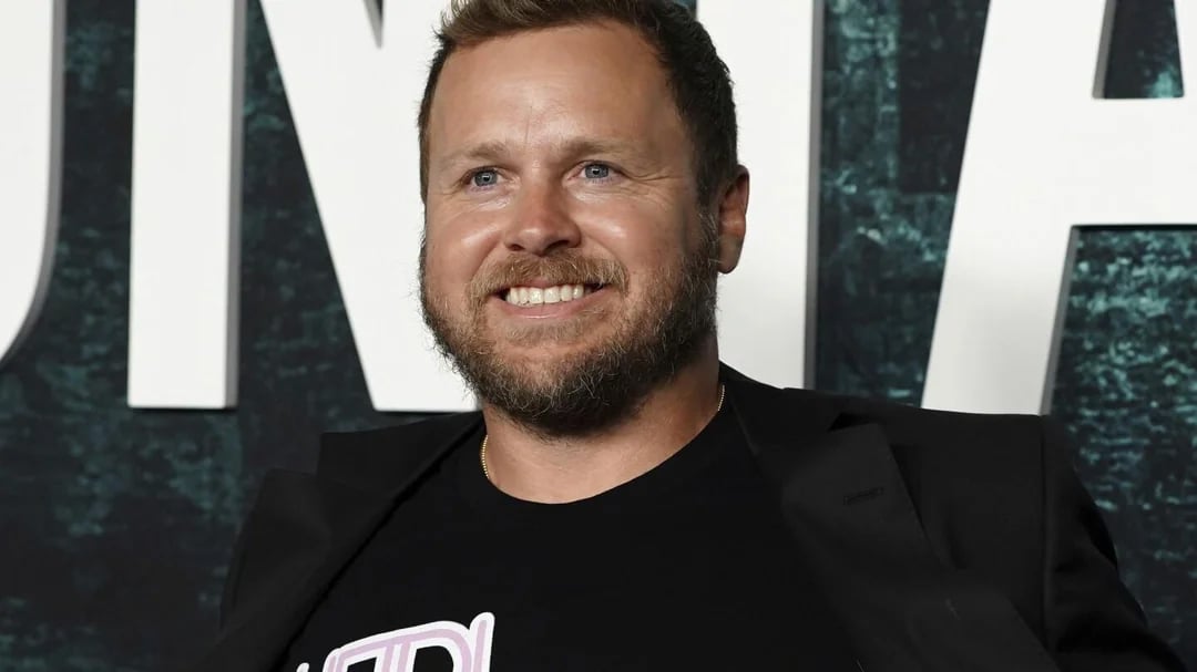 A smiling man with short hair and a beard, wearing a black T-shirt and blazer, standing in front of a backdrop with large white letters.