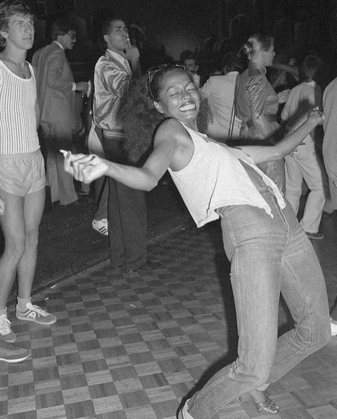 A woman with an afro and big smile dances energetically on a checkered floor at a crowded indoor party. Other people in retro clothing stand and mingle in the background. The image is in black and white.