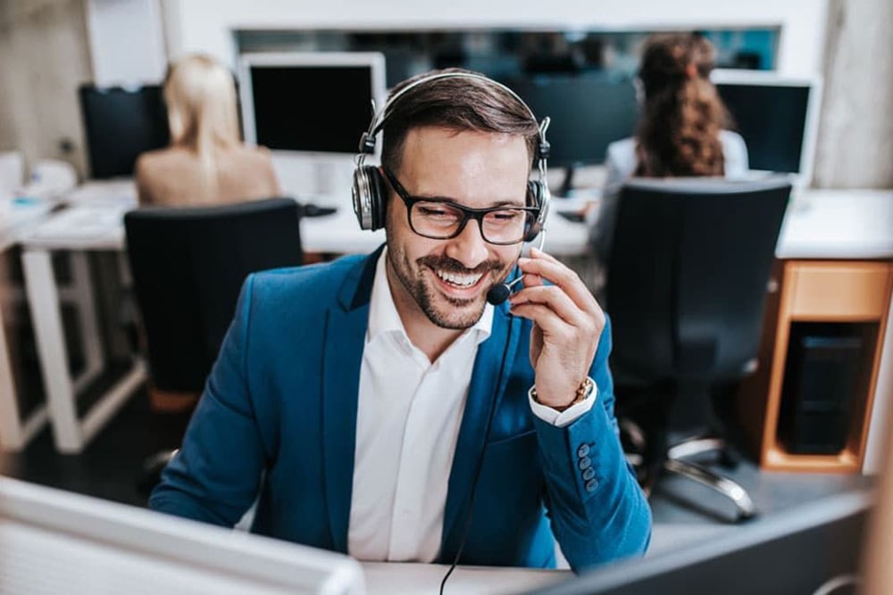 A smiling man wearing glasses and a blue blazer sits at a desk, talking into a headset microphone at an office call center, with two coworkers working at computers in the background.