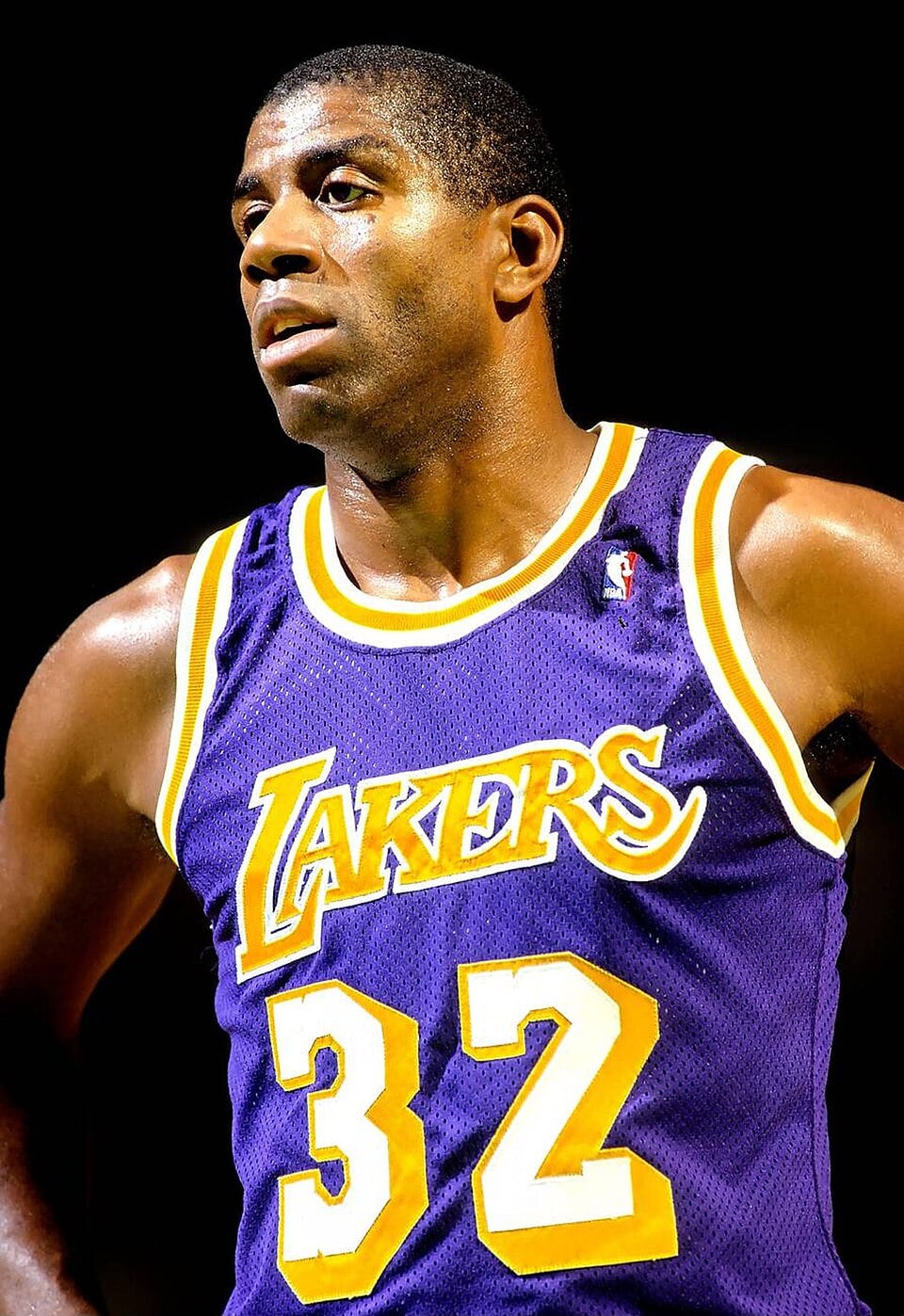 A basketball player wearing a purple and gold Lakers jersey with the number 32 stands with his hands on his hips, looking off to the side under bright lighting.