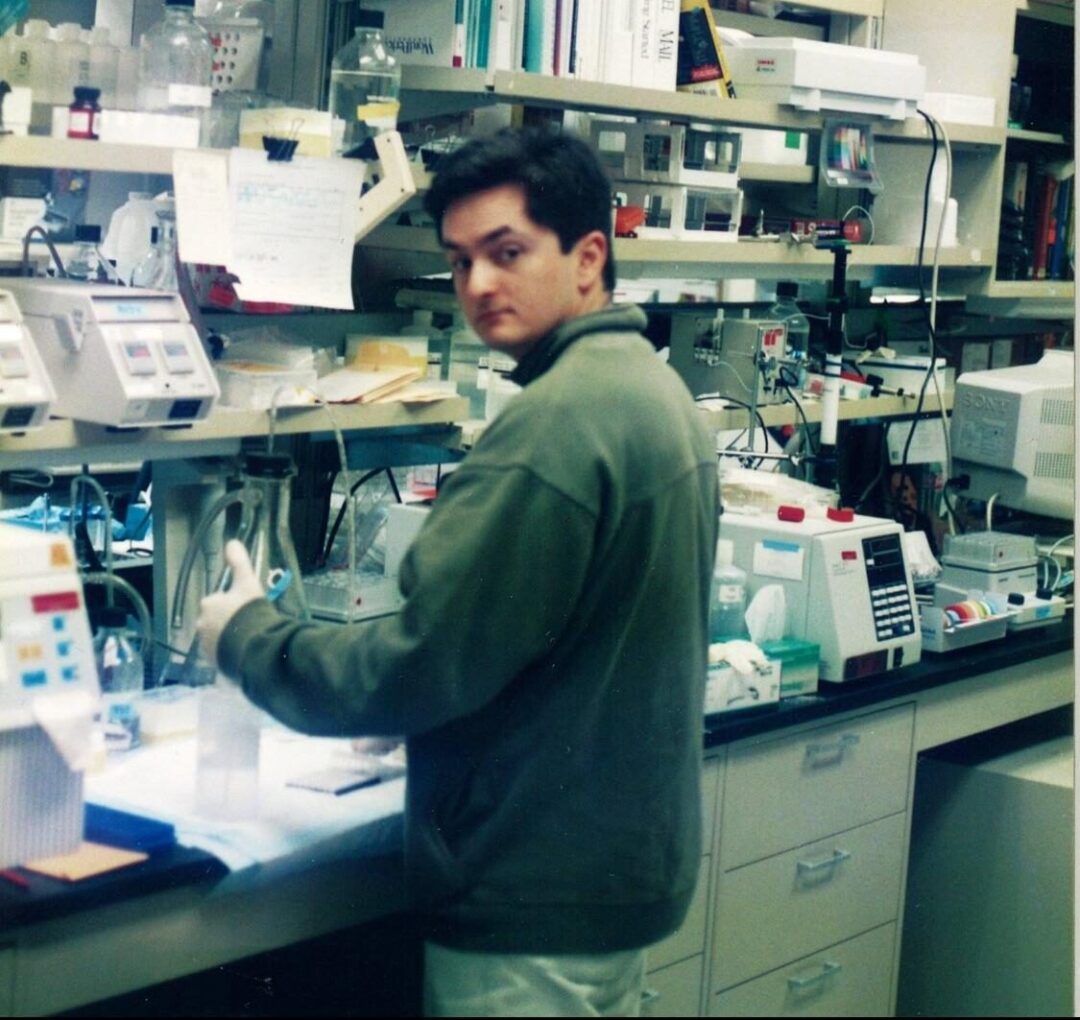 A person wearing a jacket and gloves stands at a cluttered laboratory bench with scientific equipment, bottles, and papers, looking over their shoulder toward the camera. Shelves with lab supplies are visible in the background.