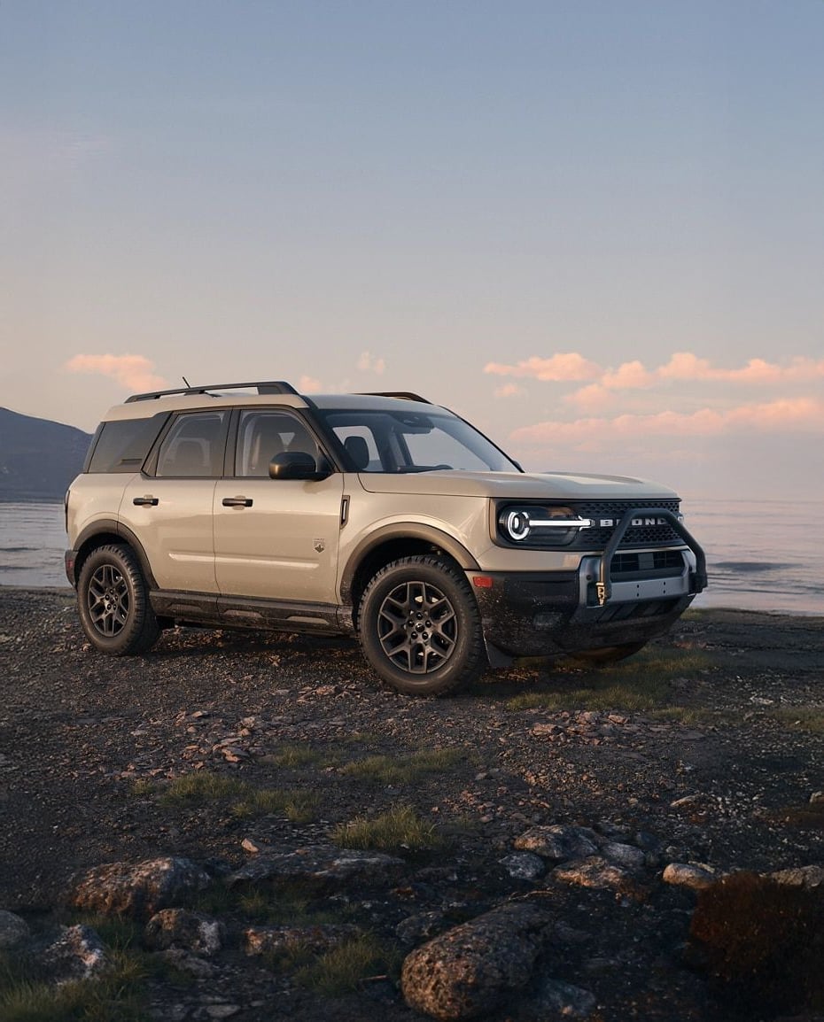 A beige Ford Bronco Sport SUV parked on rocky terrain near a body of water, with mountains and a pastel sky in the background.