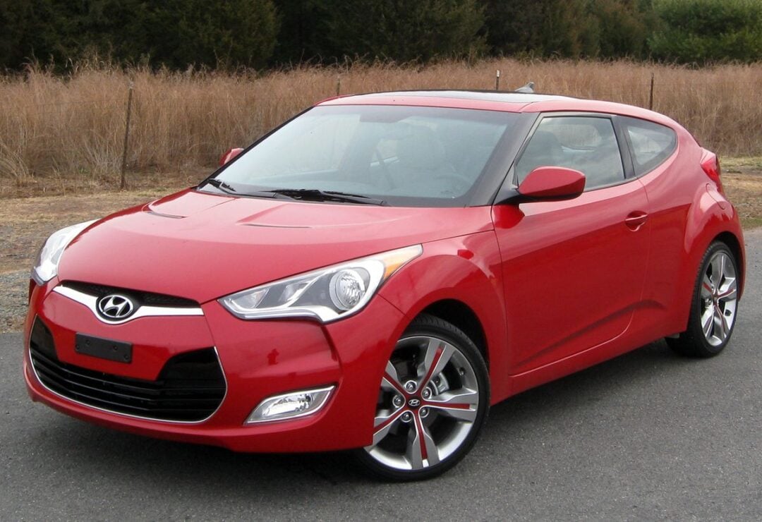 A red Hyundai Veloster hatchback is parked on a paved road beside tall dry grass, viewed from the front driver’s side. The car has sporty styling with sleek headlights and silver alloy wheels.