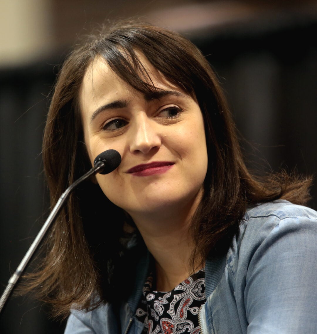 A woman with shoulder-length brown hair smiles slightly while sitting at a table with a microphone in front of her. She is wearing a light blue jacket and a patterned top, with a blurred dark background behind her.