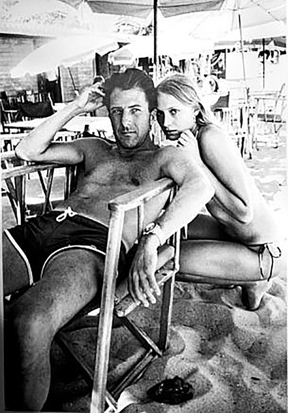 A man in swim trunks sits in a wooden chair on a sandy beach, while a woman in a bikini kneels closely beside him. Both appear relaxed under large beach umbrellas, with tables and chairs in the background.