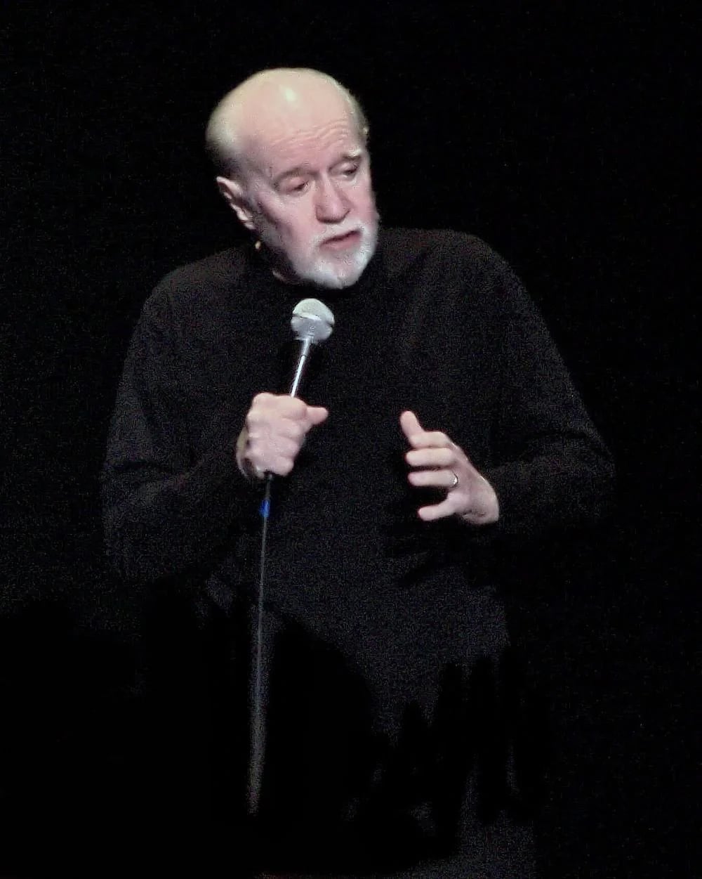A man with a white beard and bald head, dressed in a black long-sleeve shirt, is holding a microphone and speaking on stage against a dark background.
