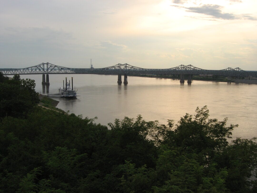 A river with a paddlewheel boat near the shore, lush greenery in the foreground, and two large steel bridges spanning the water under a cloudy sky at sunset.