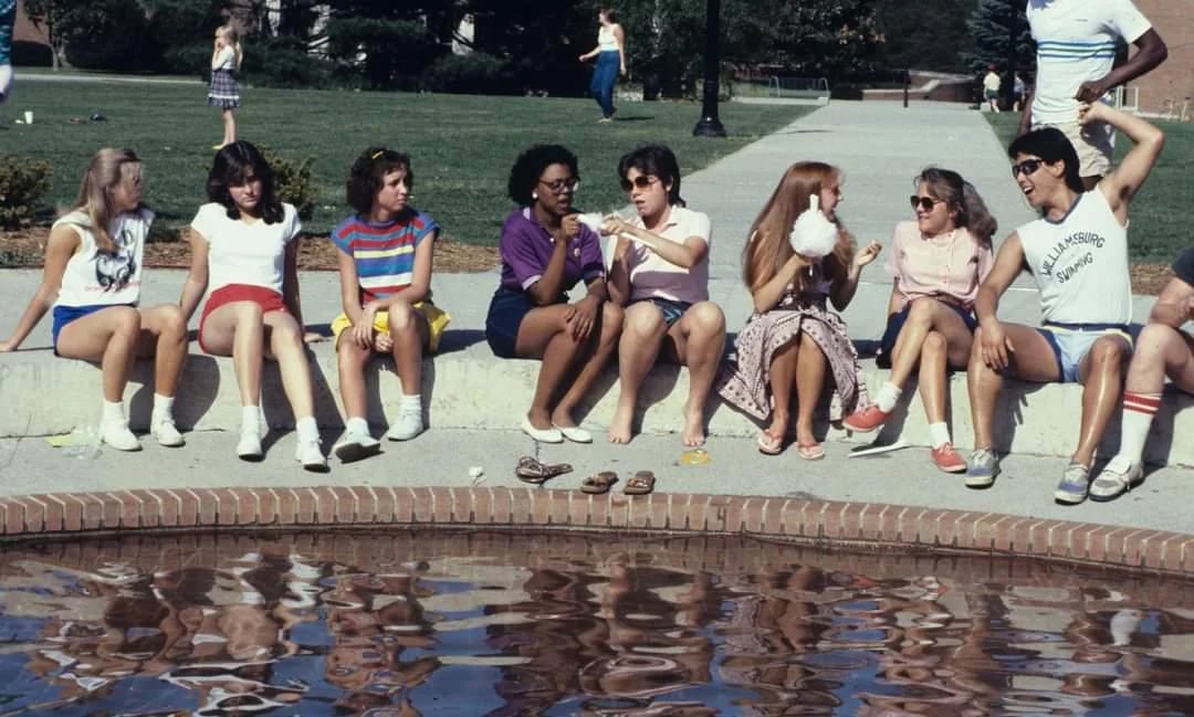 A group of young people sit on the edge of a fountain, talking and laughing together. Some have snacks or drinks, and their casual clothing suggests a warm day. Their reflections are visible in the water.