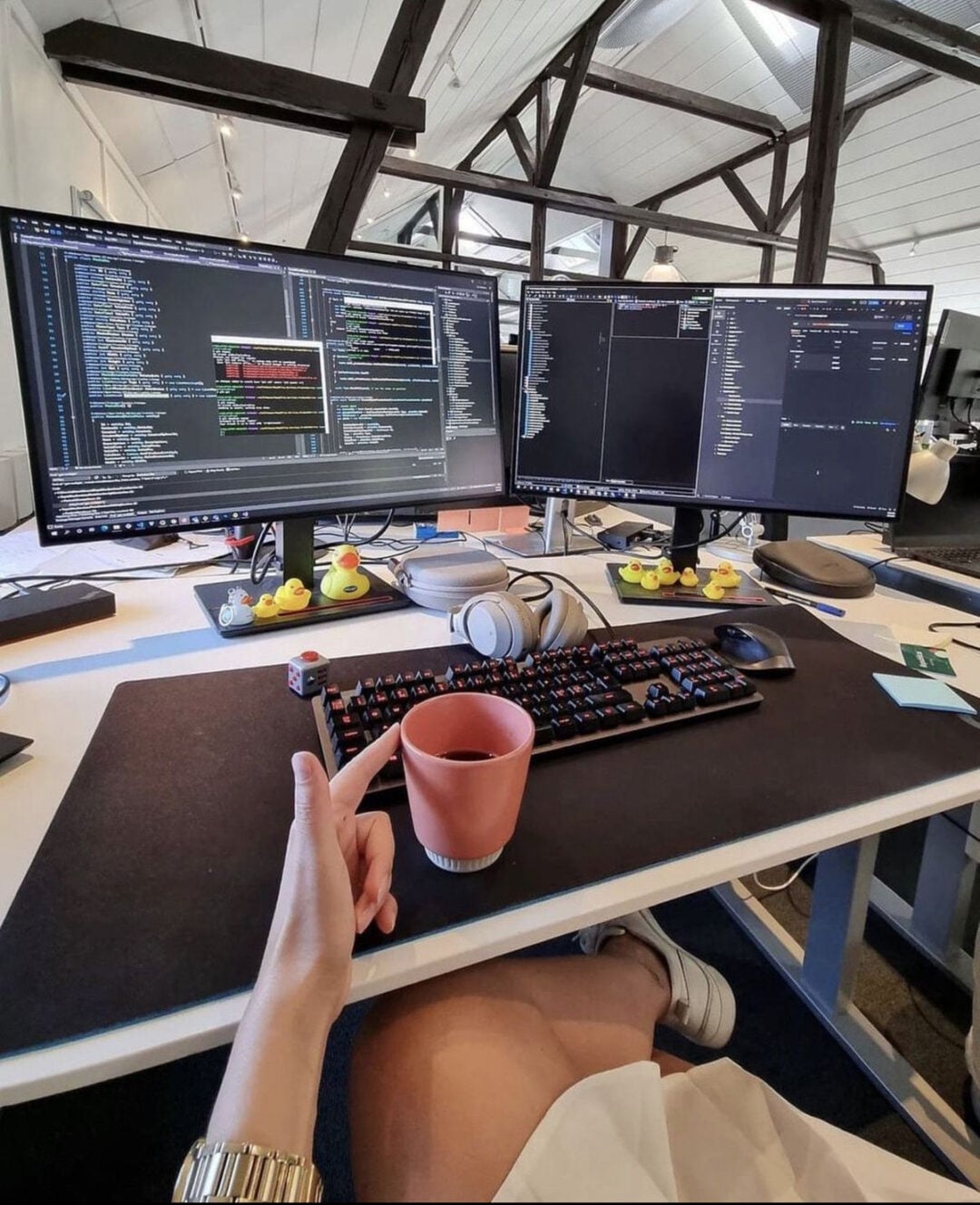 A person sits at a desk with dual monitors displaying code, a keyboard, a mouse, headphones, and a pink coffee mug. Rubber ducks line the monitor bases. The workspace features exposed wooden beams and natural light.