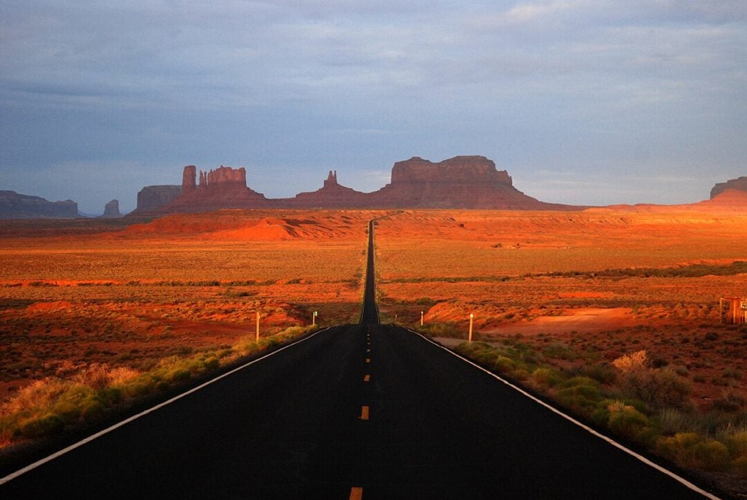 A straight, empty road stretches through a desert landscape toward tall, reddish rock formations in Monument Valley, under a partly cloudy sky with the land bathed in warm sunlight.