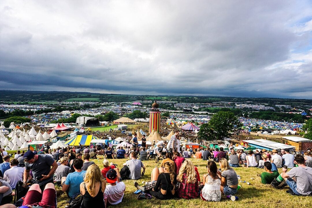 Crowds sit on a grassy hill overlooking a large, colorful festival with tents, a helter-skelter, and fairground attractions under a cloudy sky, with wide views of the event and countryside in the background.