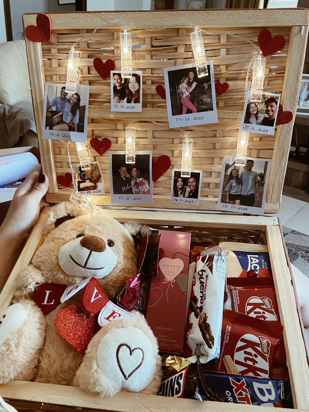 A gift box with a teddy bear, chocolates, snacks, a pink lotion, and a string of heart decorations and Polaroid photos inside the lid, each photo labeled with dates and memories.
