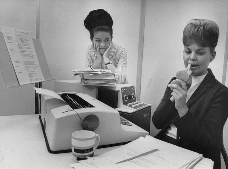 Two women in an office: one sits at a desk with a typewriter and papers, holding a compact mirror and applying lipstick; the other stands behind her, leaning on a stack of files and smiling.