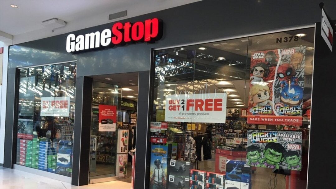 GameStop storefront in a mall with large glass windows; promotional signs and Mighty Muggs collectible figures are displayed in the window. The entrance is open and the store interior is partially visible.
