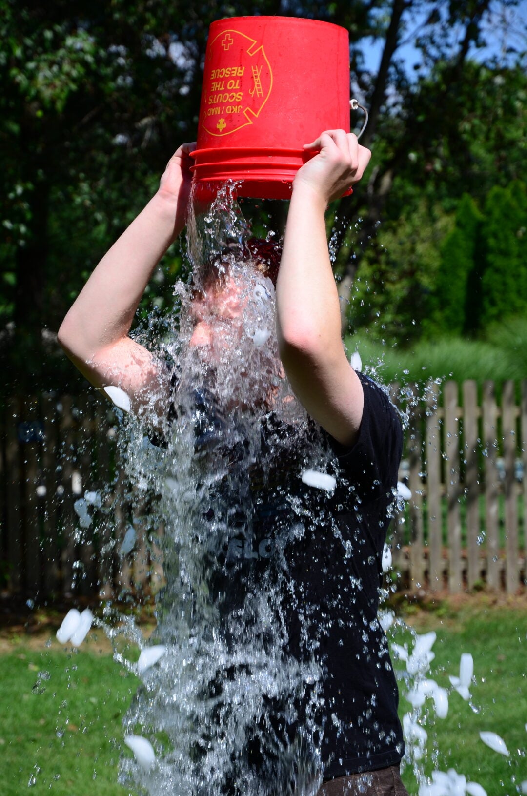 A person outdoors pours a red bucket of ice water over their head, with water splashing down and ice cubes visible. The background features a wooden fence, green grass, and trees.