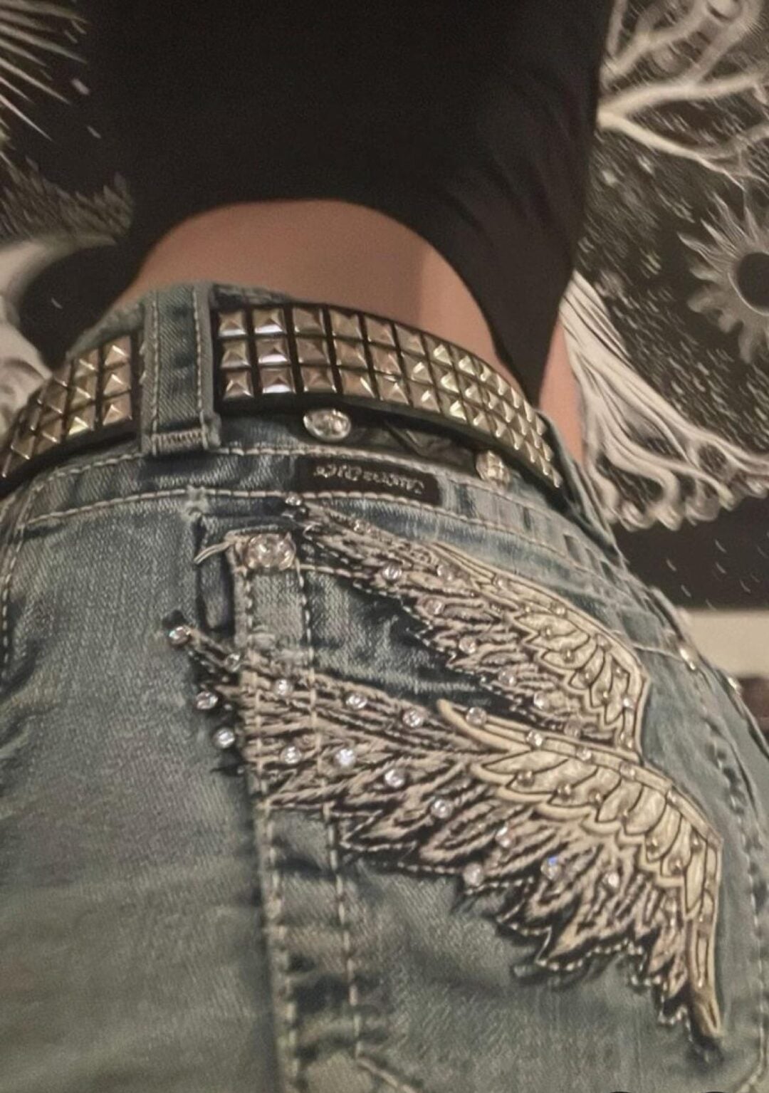 Close-up of a person wearing light blue jeans with an embellished wing design on the pocket, a studded belt, and a black shirt. The background shows a black and white patterned surface.