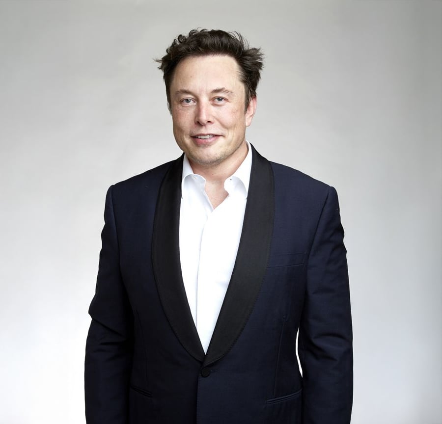 A man wearing a black tuxedo with a white shirt stands against a plain, light-colored background, smiling slightly at the camera.