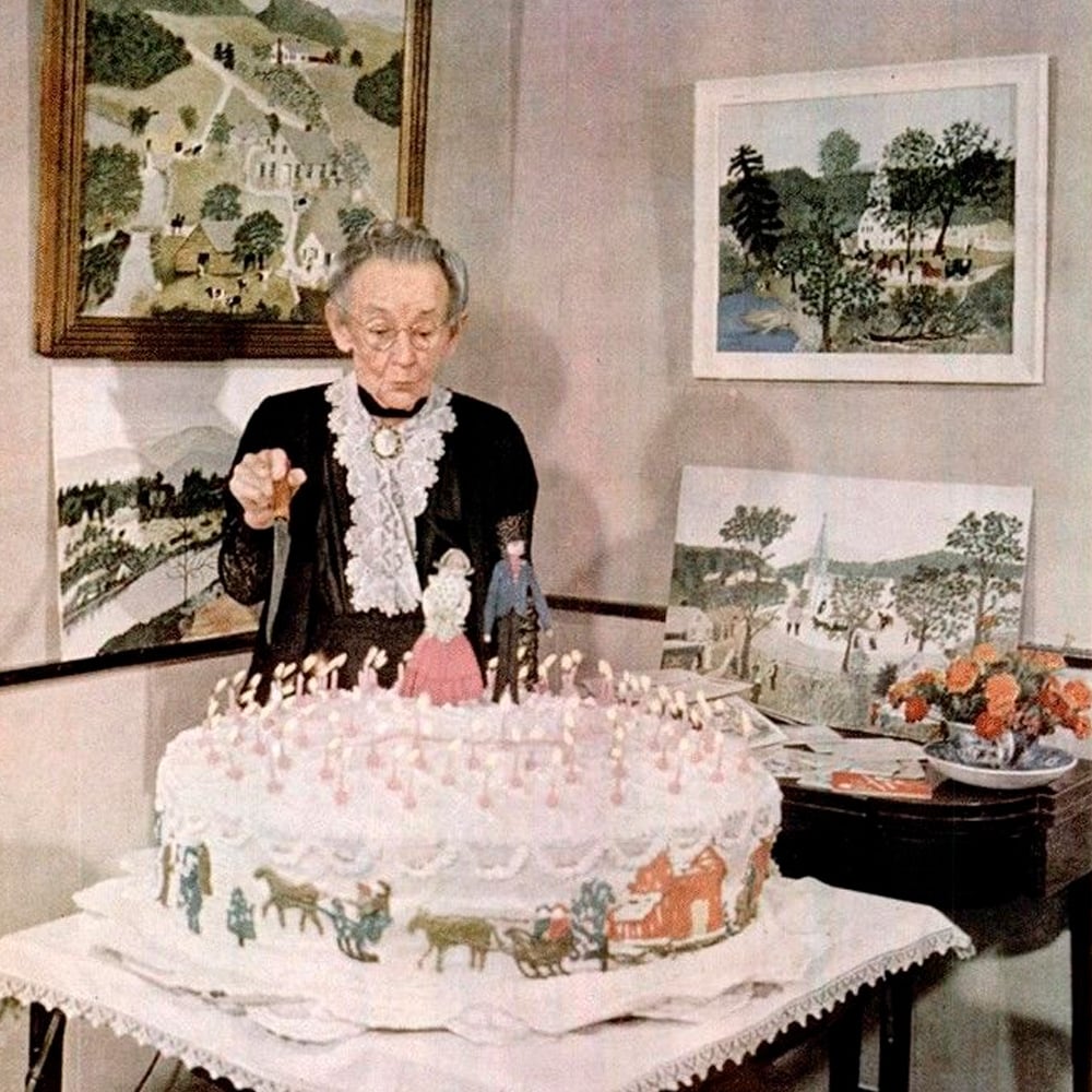 An elderly woman in a black dress stands behind a large, decorated birthday cake with many candles. Folk art paintings are displayed on the wall and table behind her, along with a vase of flowers.