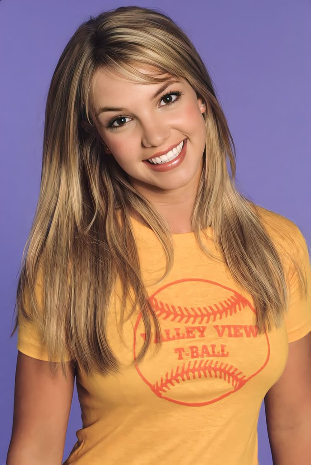 A young woman with long blonde hair smiles at the camera. She is wearing a yellow t-shirt with a red baseball graphic and the words "VALLEY VIEW T-BALL" printed on it. The background is solid purple.