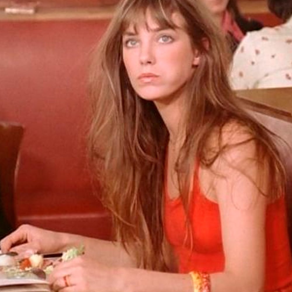 A woman with long brown hair wearing a sleeveless red top sits in a booth at a restaurant, holding utensils and looking slightly upward with a neutral expression.