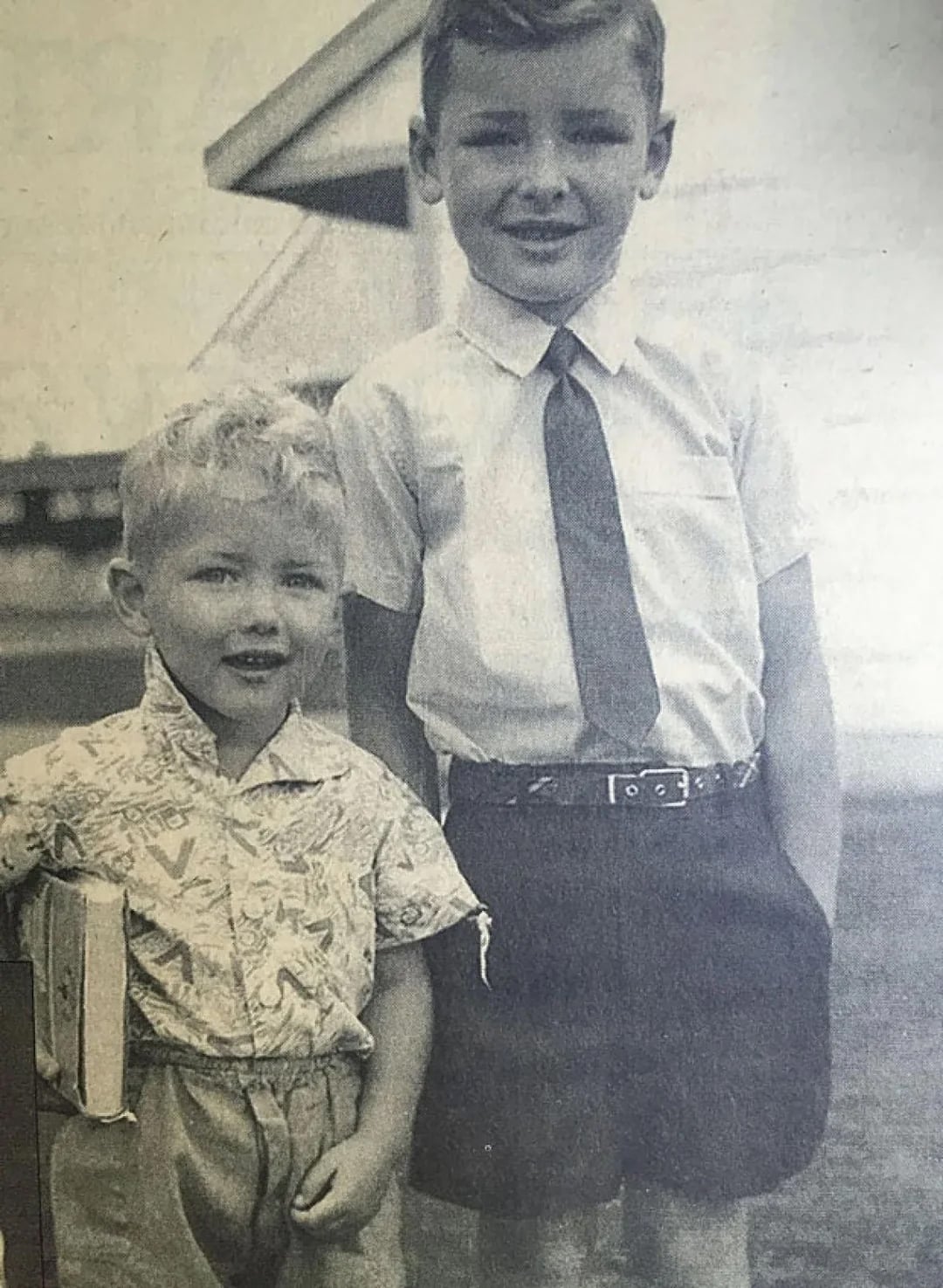 Two young boys stand outside. The older boy wears a white shirt, tie, and dark shorts, smiling at the camera. The younger boy, holding a book, wears a patterned shirt and light shorts. The photo is in black and white.