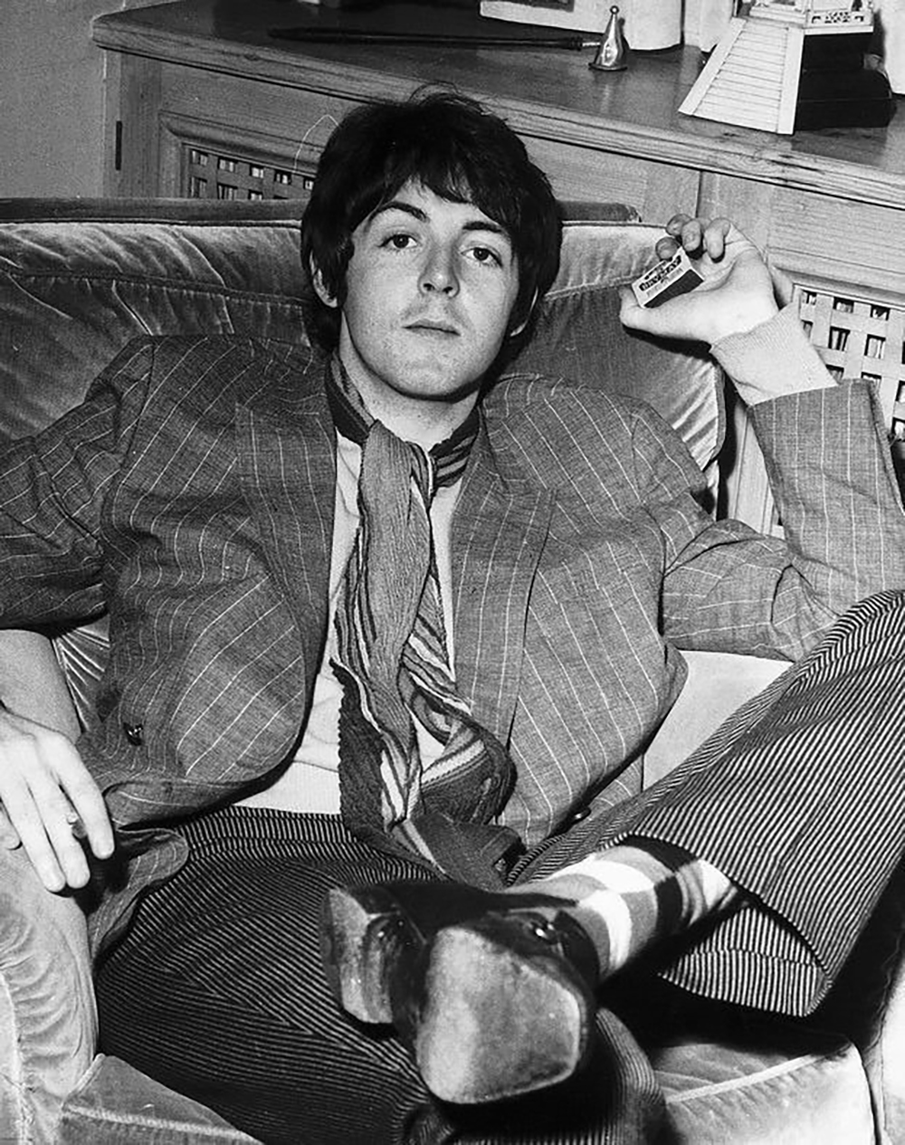 A young man in a pinstripe suit and striped scarf sits casually in an armchair, holding a small object in his raised hand, with one leg crossed over the other. The background shows a table with various items.