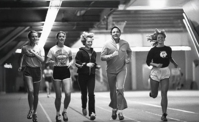Five people jog side by side on an indoor track, smiling and wearing athletic clothing. The setting is well-lit and appears energetic, with motion visible especially from the runner on the right.
