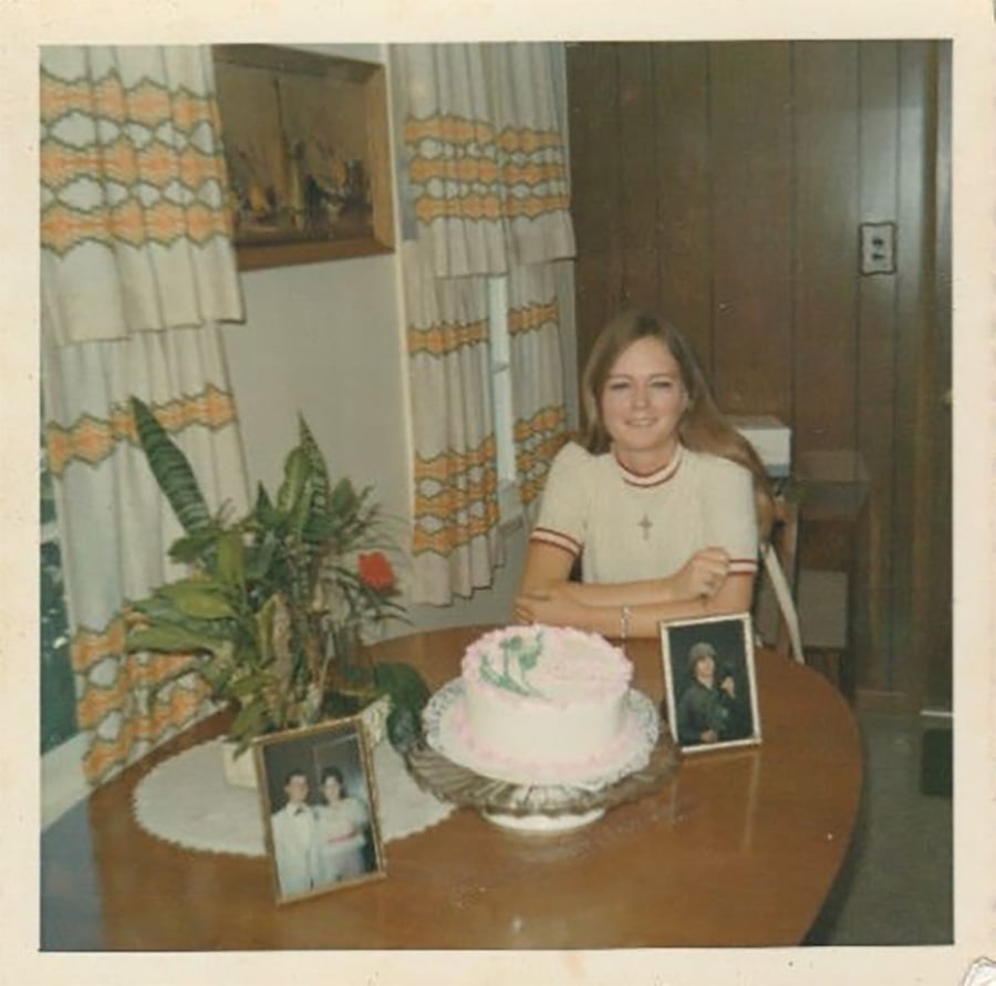 A young woman sits at a wooden table with a pink and white cake, two framed photos, and a potted plant. She is smiling, and the room has patterned curtains and wood-paneled walls.