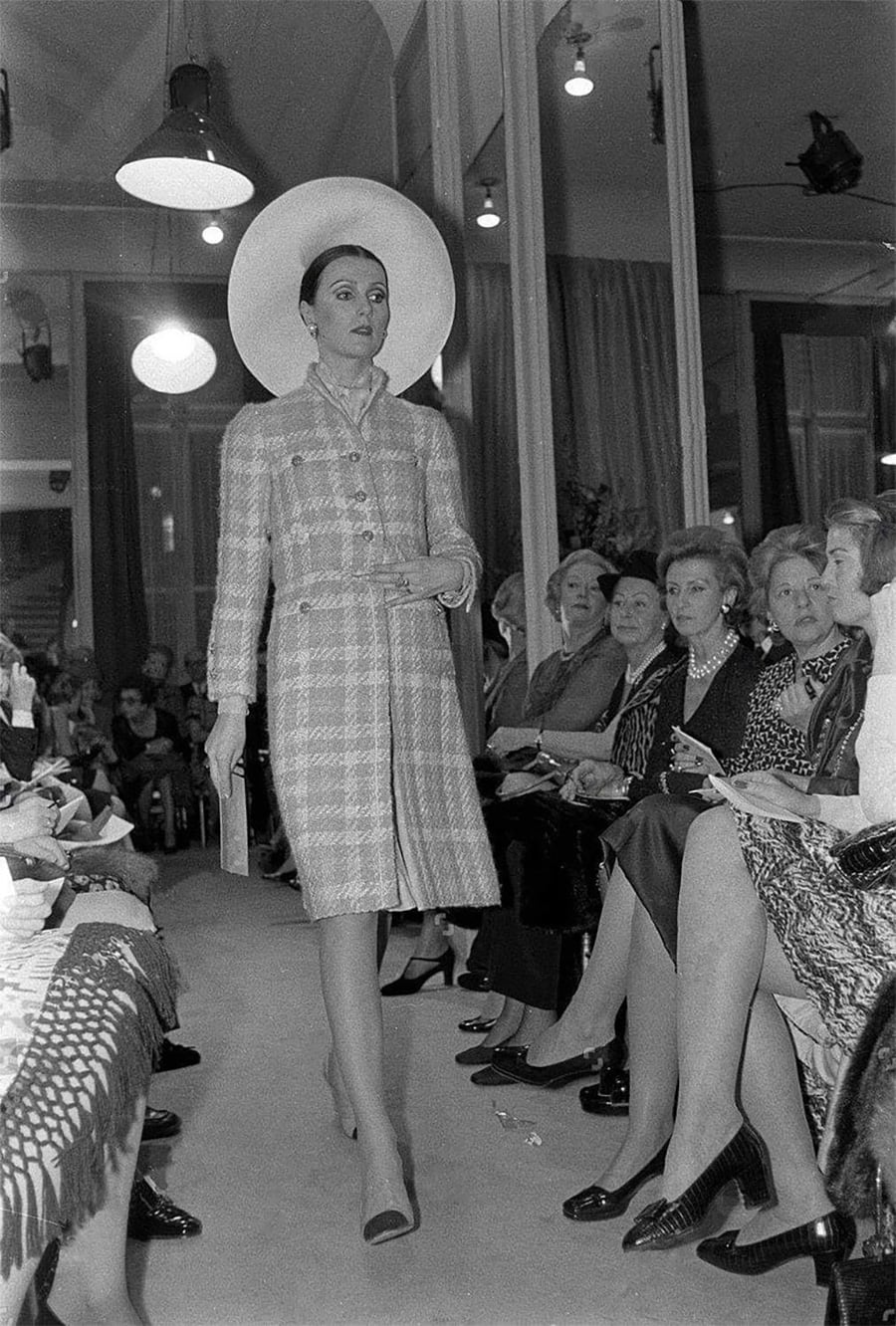 A model in a plaid coat and wide-brimmed hat walks the runway at a fashion show, surrounded by seated audience members in formal attire observing attentively.