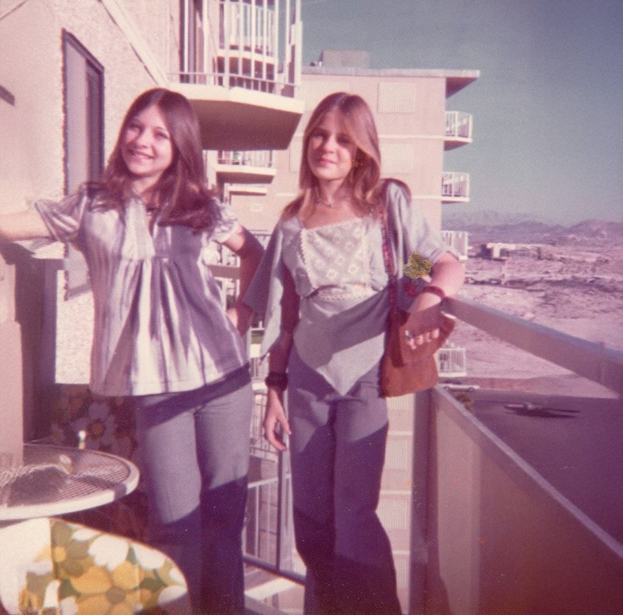 Two young women with long hair stand on a balcony, smiling. Both are wearing 1970s-style outfits with flared pants and patterned tops. Apartment buildings and a desert landscape are visible in the background.