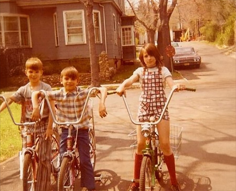 Three children with short and long hair pose on vintage bicycles on a suburban street, with a house and parked car in the background. The photo has a warm, nostalgic tone, suggesting it was taken in the 1970s.