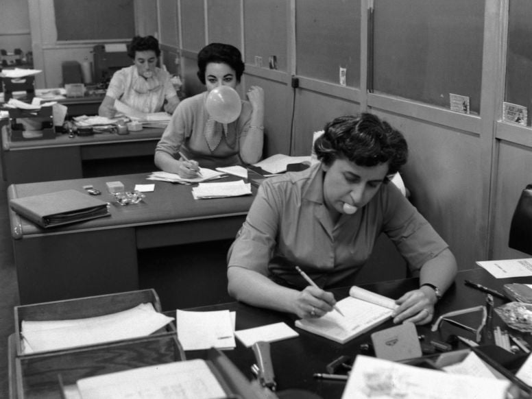 Three women are sitting at desks in an office. Two are writing or working, while the woman in the middle is blowing a bubble with chewing gum. Papers and office supplies are spread across their desks.