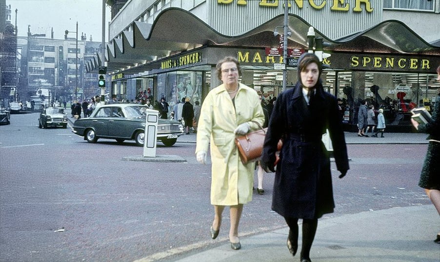Two women walk on a city street near a Marks & Spencer store. One wears a yellow coat and carries a brown bag, while the other wears a dark coat and scarf. Vintage cars and buildings are visible in the background.