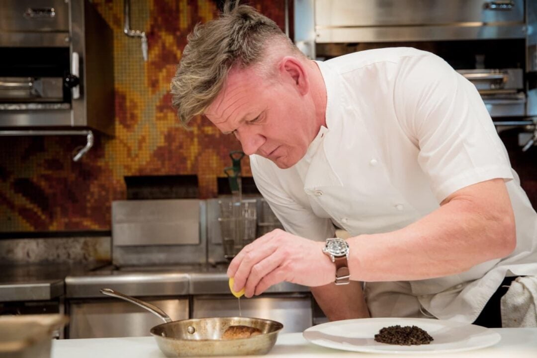 A chef in a white uniform squeezes a lemon over a pan on a kitchen counter, focused on his cooking. A white plate with food is in front of him, and there are stainless steel appliances in the background.