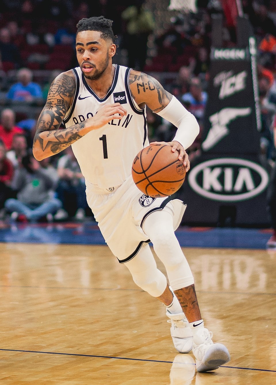 A basketball player in a Brooklyn Nets uniform dribbles the ball on a basketball court during a game, with spectators and advertisement boards visible in the background.