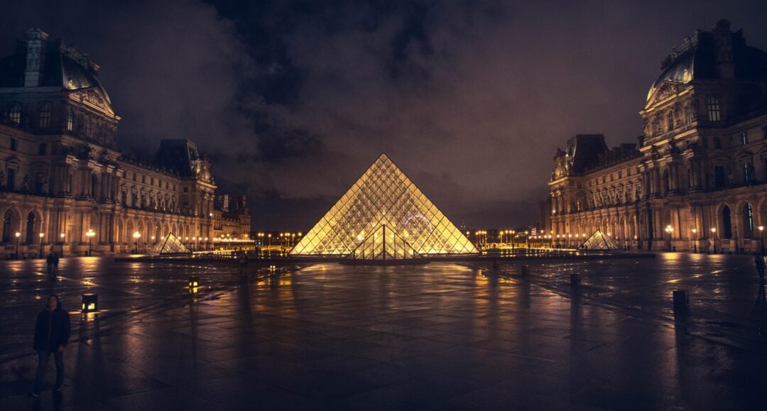 The Louvre Pyramid in Paris is illuminated at night, surrounded by the historic museum buildings. The wet ground reflects the lights, and a few people are visible in the spacious plaza.