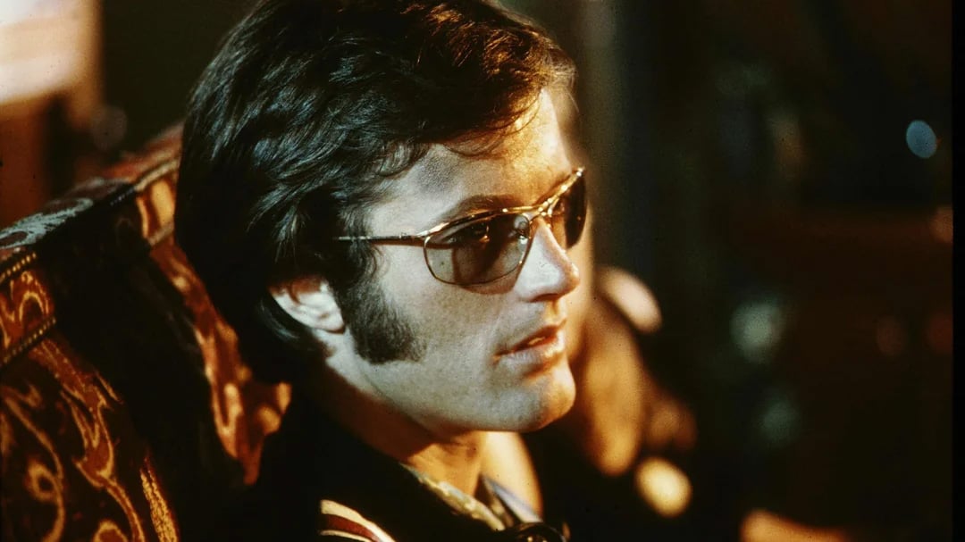 A man with dark hair and sideburns wearing aviator sunglasses sits on a patterned chair, gazing thoughtfully to the side in a softly lit setting.