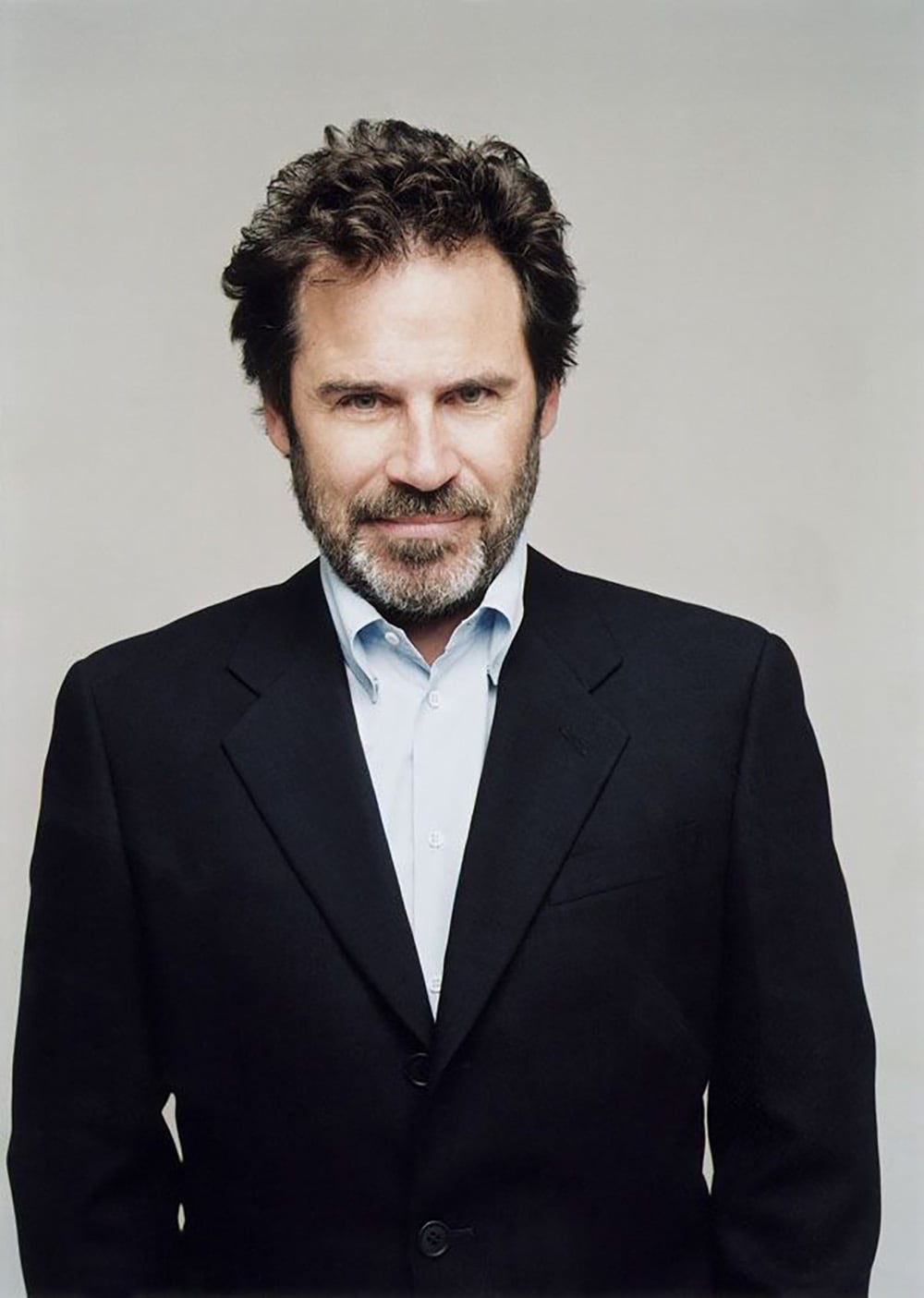 A man with dark, wavy hair and a beard is wearing a black suit jacket over a light blue shirt. He is standing against a plain, light-colored background and looking directly at the camera with a slight smile.