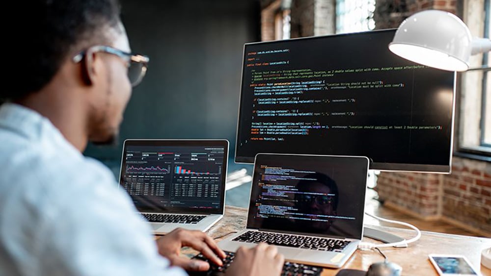 A person sits at a desk working on a laptop with code on the screen, while two monitors in front display data charts and programming code in a modern office setting.