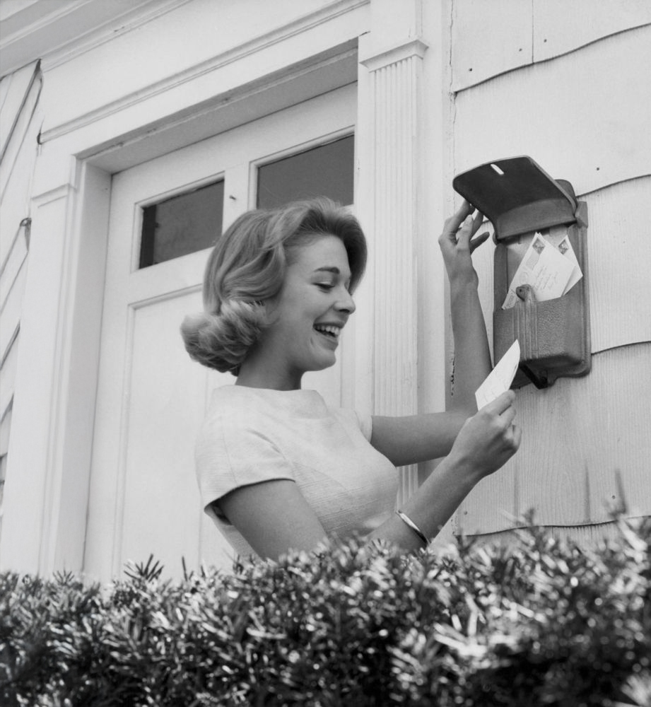 A woman smiles as she stands outside by a house, taking mail from a wall-mounted mailbox. She holds an envelope in one hand and opens the mailbox lid with the other. Shrubs are visible in the foreground.